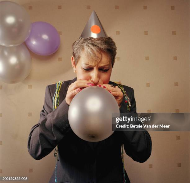 woman wearing party hat, blowing into balloon, eyes closed - silver balloon imagens e fotografias de stock