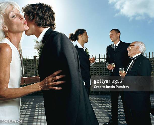 senior woman greeting young man with kiss, three men in background - cheek stock pictures, royalty-free photos & images