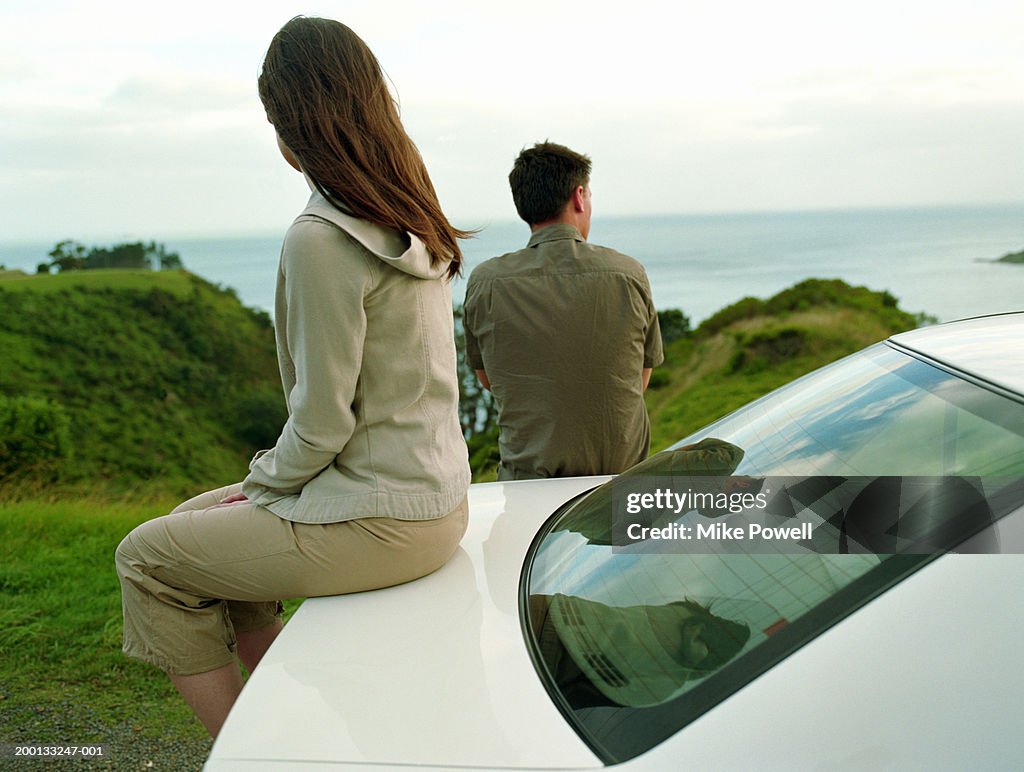 Young couple looking at sea, woman sitting on trunk of car