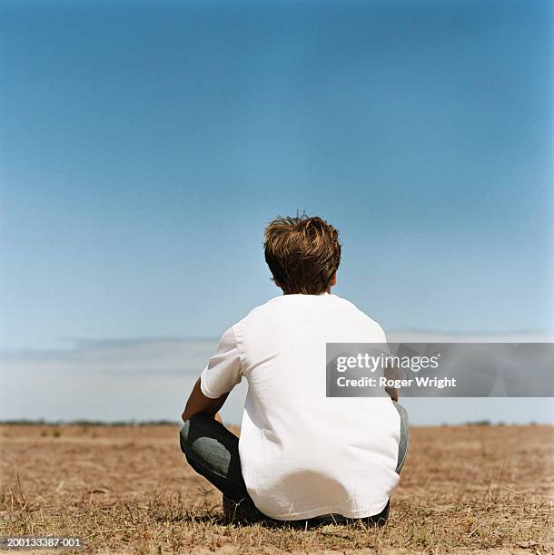 young man sitting cross legged in field, rear view - schneidersitz stock-fotos und bilder