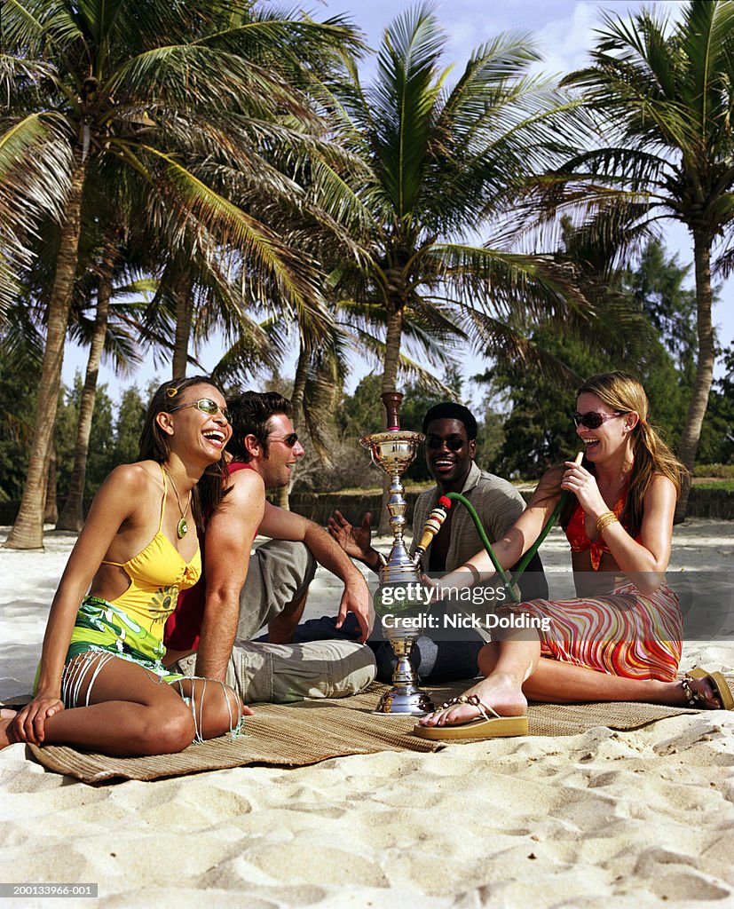 Four young people on beach near palm trees, woman holding hooka pipe