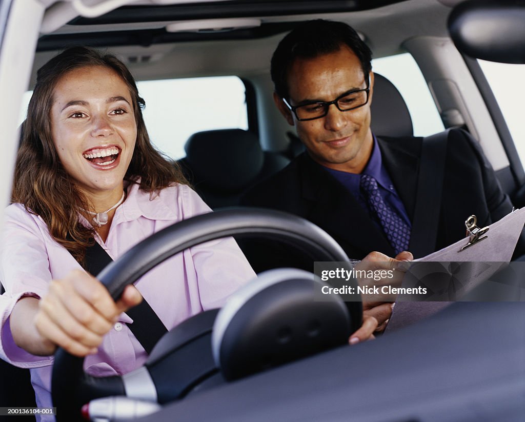 Woman driving car smiling, man in passenger seat with clip-board