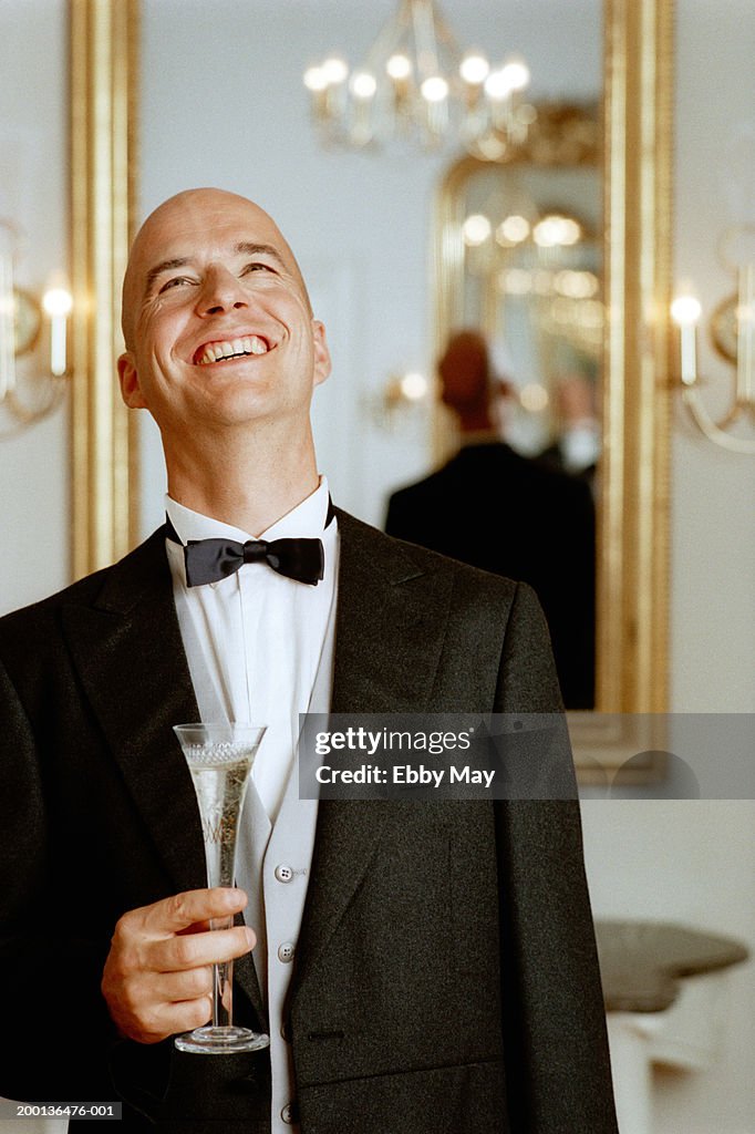 Man wearing tuxedo, holding champagne flute looking up, smiling