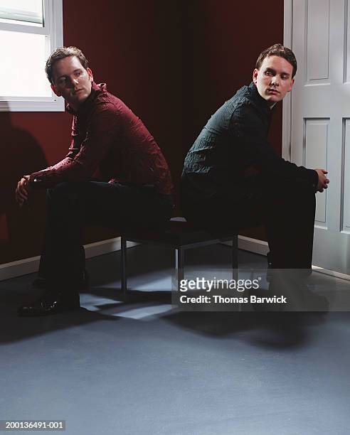 two young men back to back on chairs, looking at each other - sibling conflict stock pictures, royalty-free photos & images