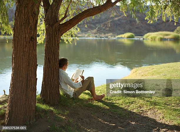 man reading book under tree by river - bare feet male tree stock pictures, royalty-free photos & images