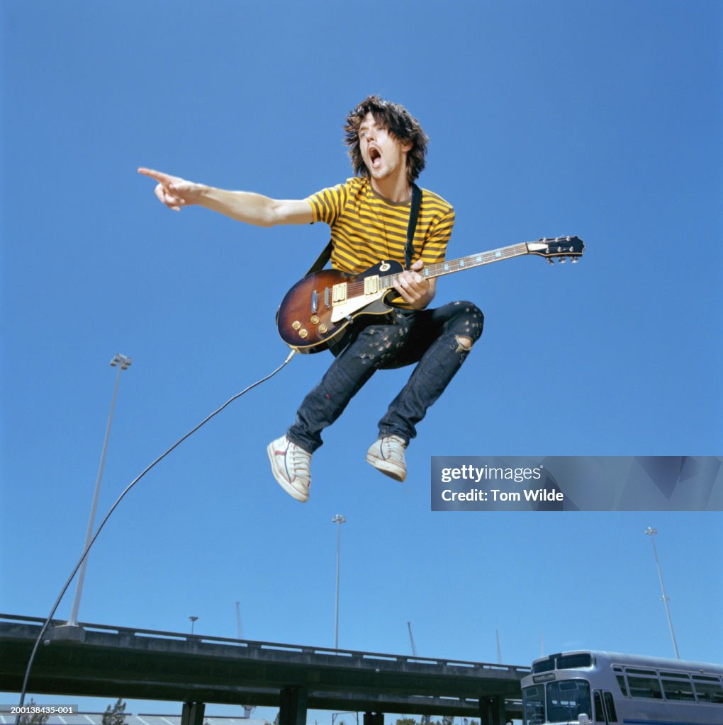 Young man with guitar leaping in air outdoors, low angle view