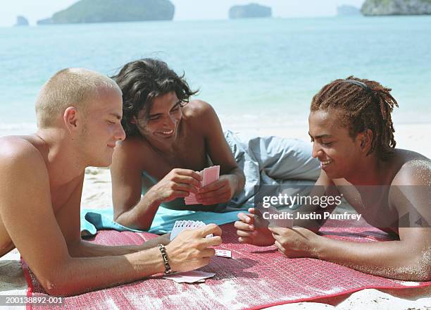 three young men playing card game on beach, leaning on beach mat - beach mat stock pictures, royalty-free photos & images