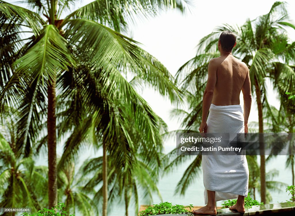 Man with towel around waist, standing on wall facing palms, rear view
