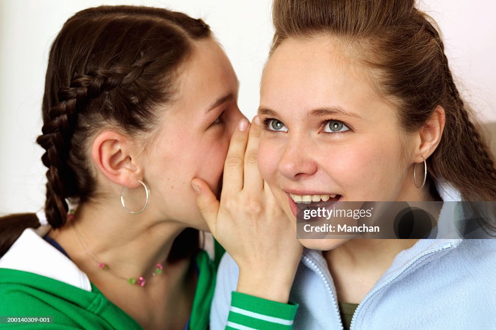 Two teenage girls (13-15), one whispering into another's ear, close up