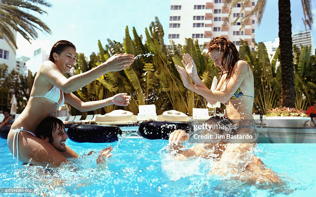 Two women on men's shoulders in swimming pool play fighting, side view