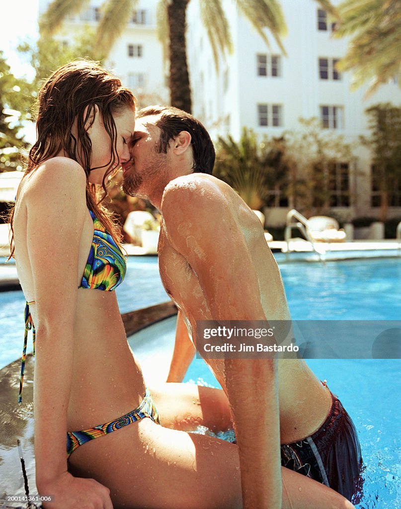 Young man kissing woman sitting on swimming pool edge, side view