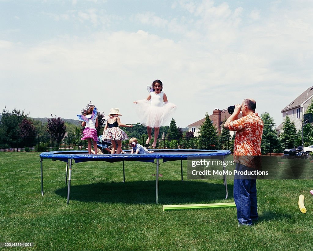 Father filming children (2-12) playing on trampoline on lawn