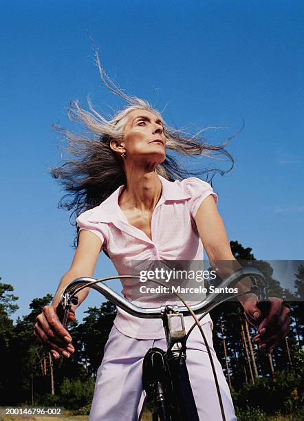 mature woman riding bicycle, hair blowing in wind, low angle view - stoneplus4 stock pictures, royalty-free photos & images