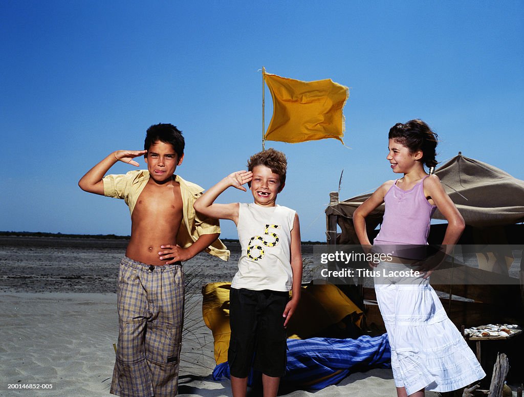 Girl and two boys (7-10) by makeshift shelter on beach, boys saluting