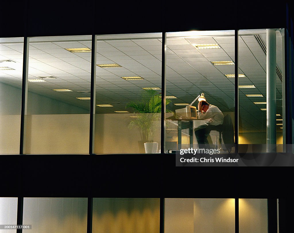 Man at desk in office, head in hands, view through window, night