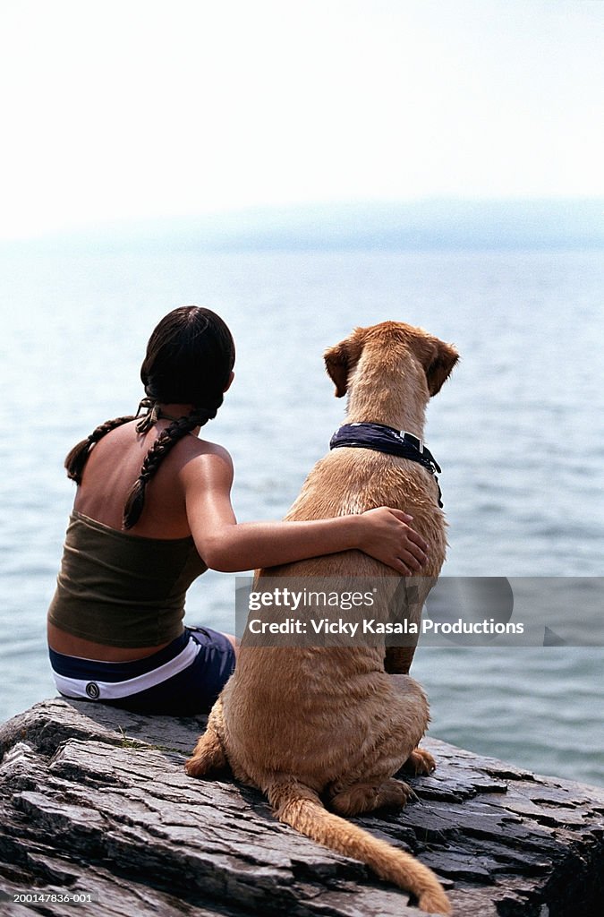 Young woman and golden lab sitting on rock, looking at lake, rear view