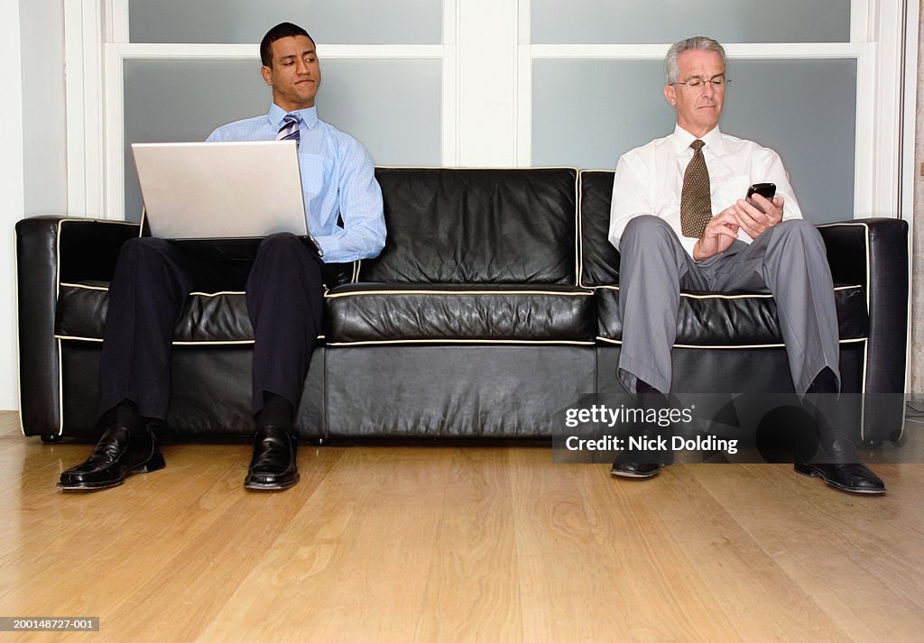Two men on sofa, one with laptop, another holding palm top computer