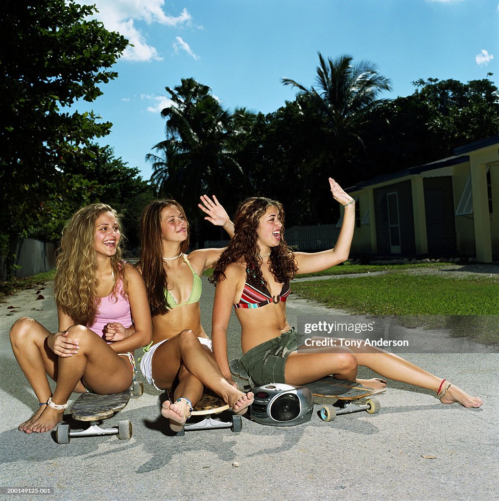 Three teenage girls (16-18) sitting on skateboards, waving