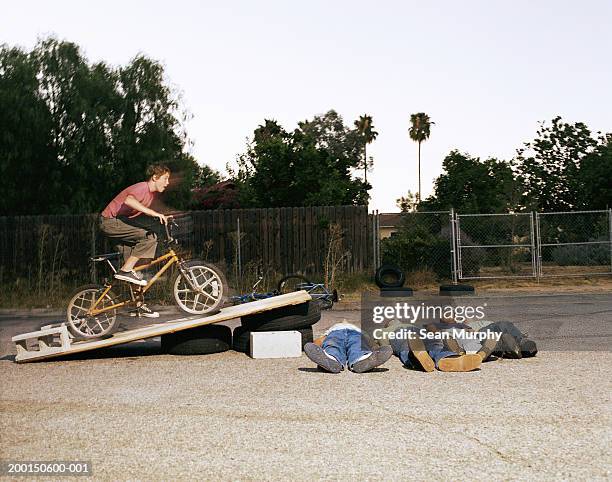 boy (11-15) riding bicycle off jump over friends (blurred motion) - bicycle stunt stock pictures, royalty-free photos & images