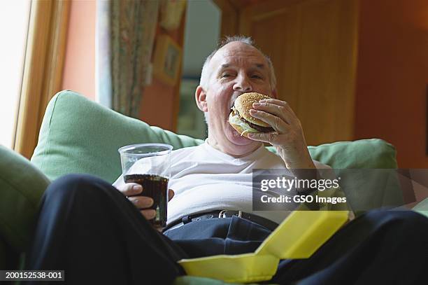 senior man on sofa, eating hamburger - unhealthy eating 個照片及圖片檔