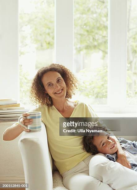 mother and son (5-7) relaxing on sofa, smiling, portrait - family on couch with mugs stock-fotos und bilder