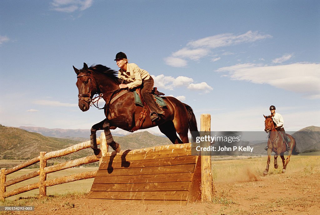 Horsewoman and horseman jumping cross-country fence , side view