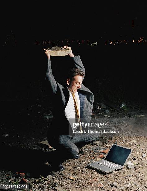businessman about to smash laptop with rock at deserted location - technofobie stockfoto's en -beelden