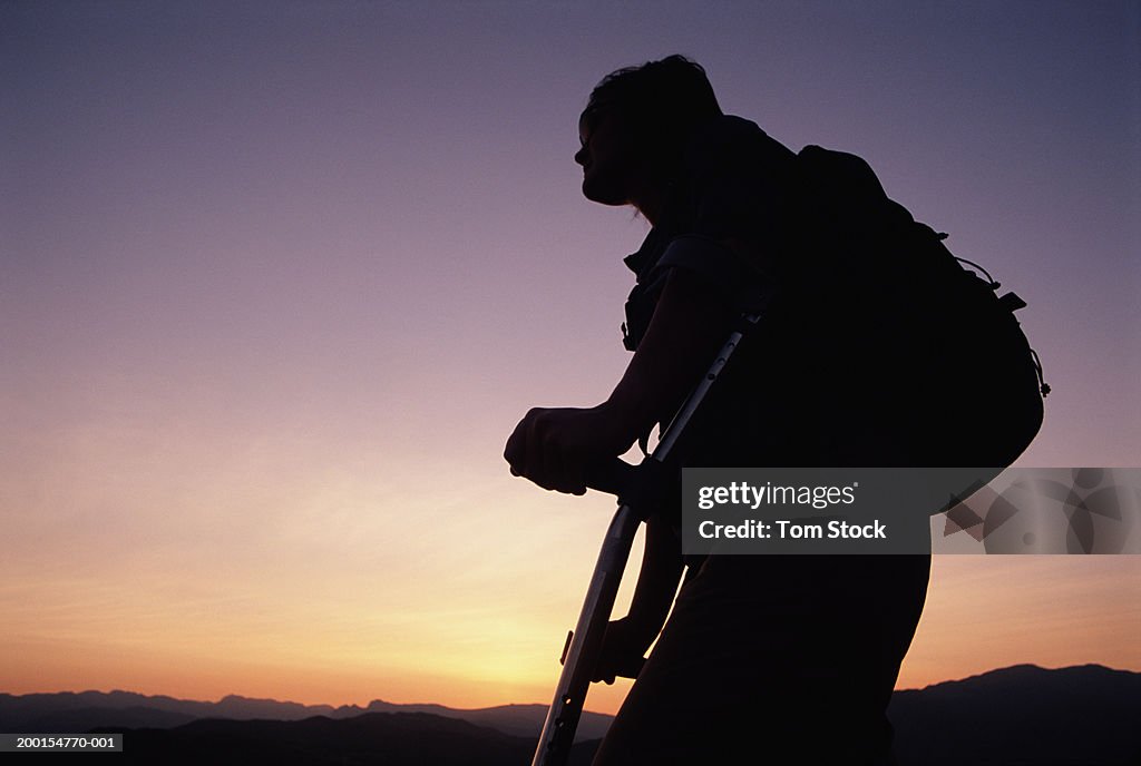 Woman with rucksack using crutches in rural environment, sunset
