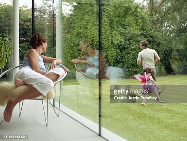 woman looking out of window at father and daughter running in garden - mother running stock-fotos und bilder