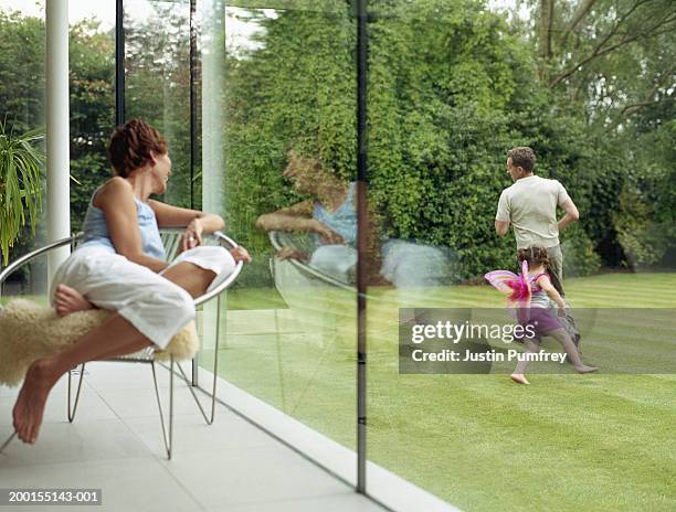 woman looking out of window at father and daughter running in garden - windows foto e immagini stock