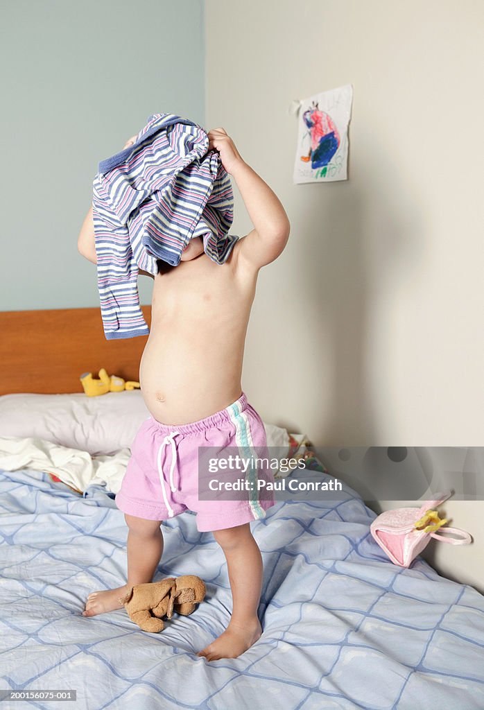 Girl (2-4) standing on bed, pulling shirt over head