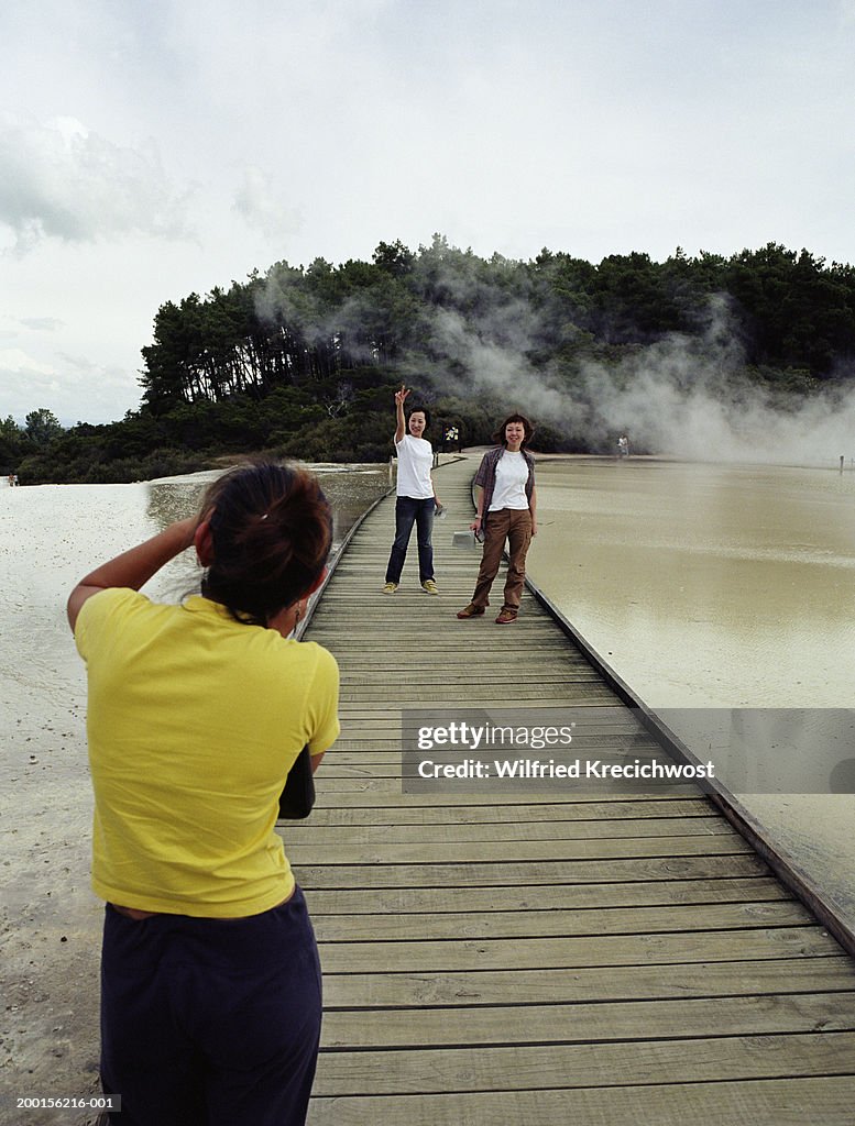 Woman taking photograph of friends on wooden path over thermal pool