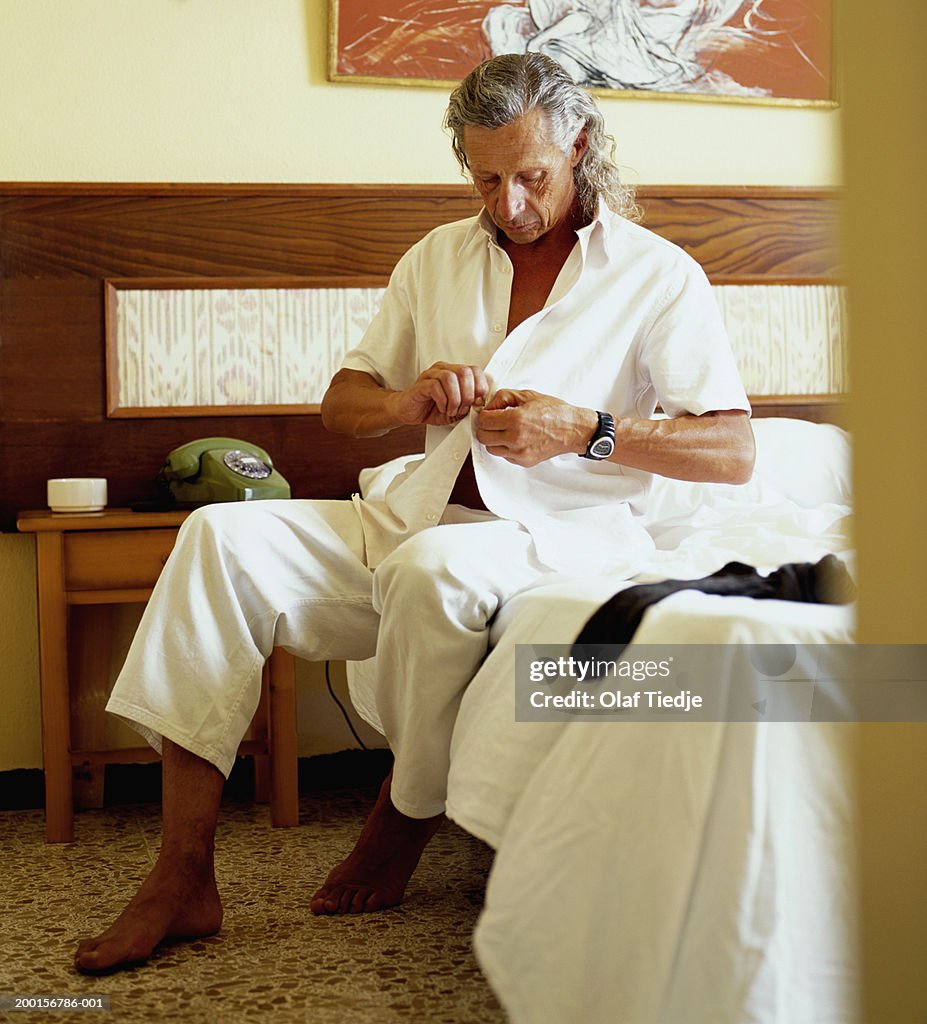 Mature man sitting on bed, buttoning shirt