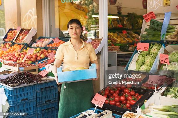 woman standing outside greengrocers, holding box of melons, portrait - grocer stock-fotos und bilder