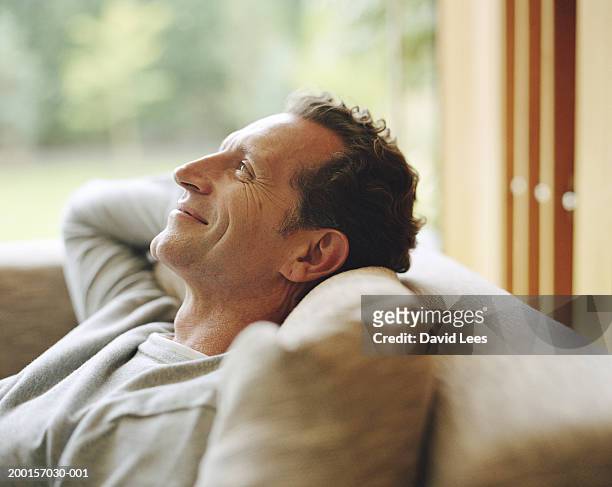 mature man relaxing on sofa, hands behind head, smiling, profile - homme content chez lui photos et images de collection