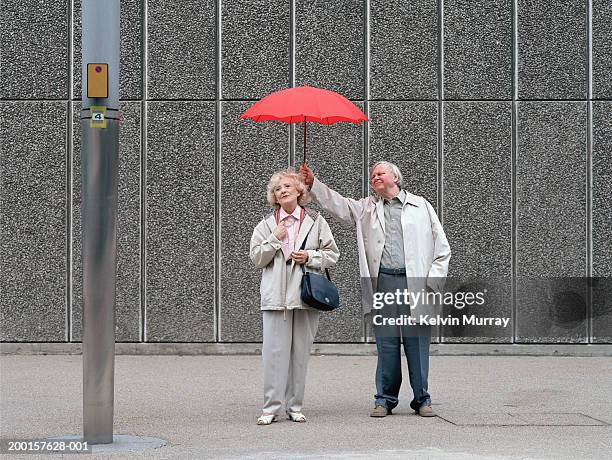senior man holding red umbrella over woman, standing on pavement - uitdelen stockfoto's en -beelden