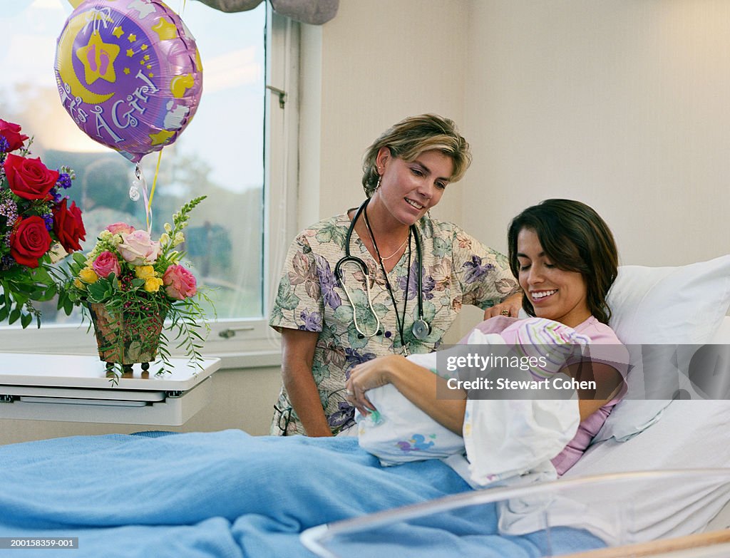 Mother holding newborn baby in hospital room, nurse looking on