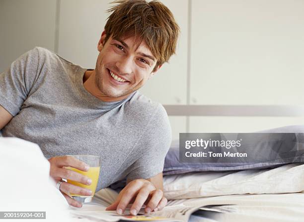 young man lying on bed by magazine, holding drink, smiling, portrait - glass magazine stock-fotos und bilder