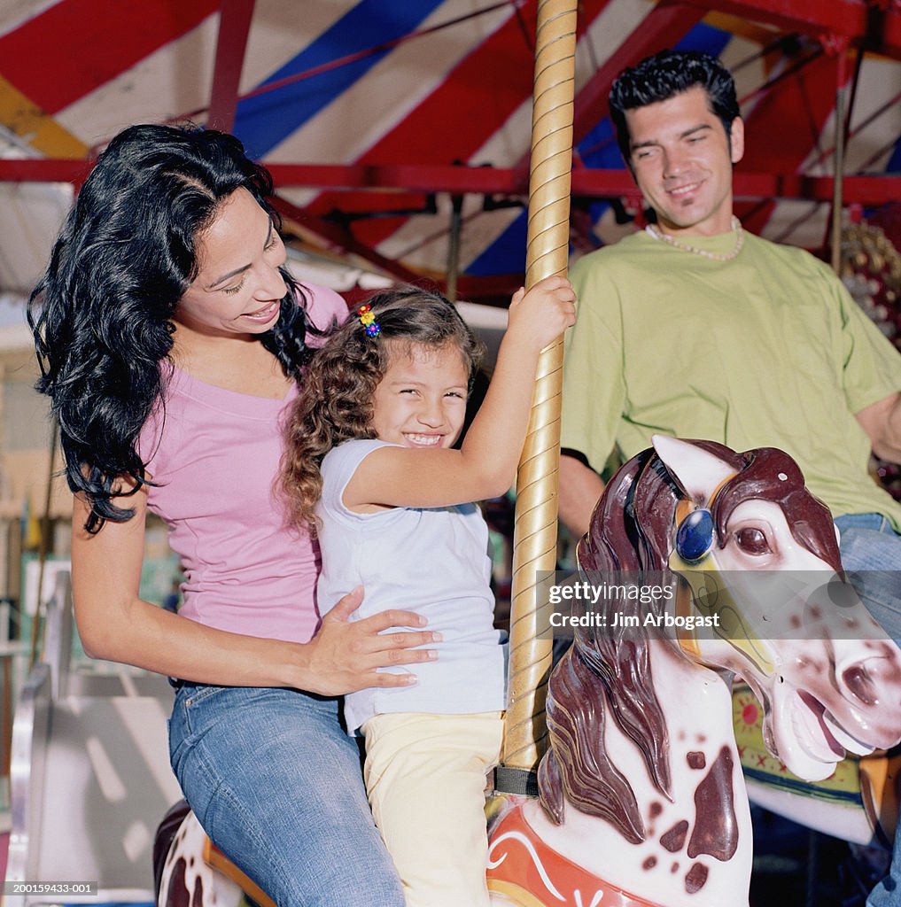 Parents and daughter (3-5) riding on carousel, smiling