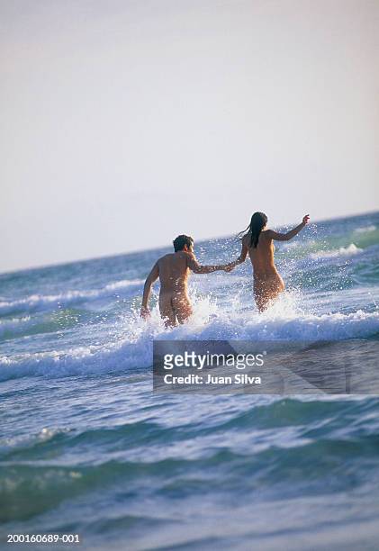 naked young couple running in surf, holding hands, rear view - nudity foto e immagini stock