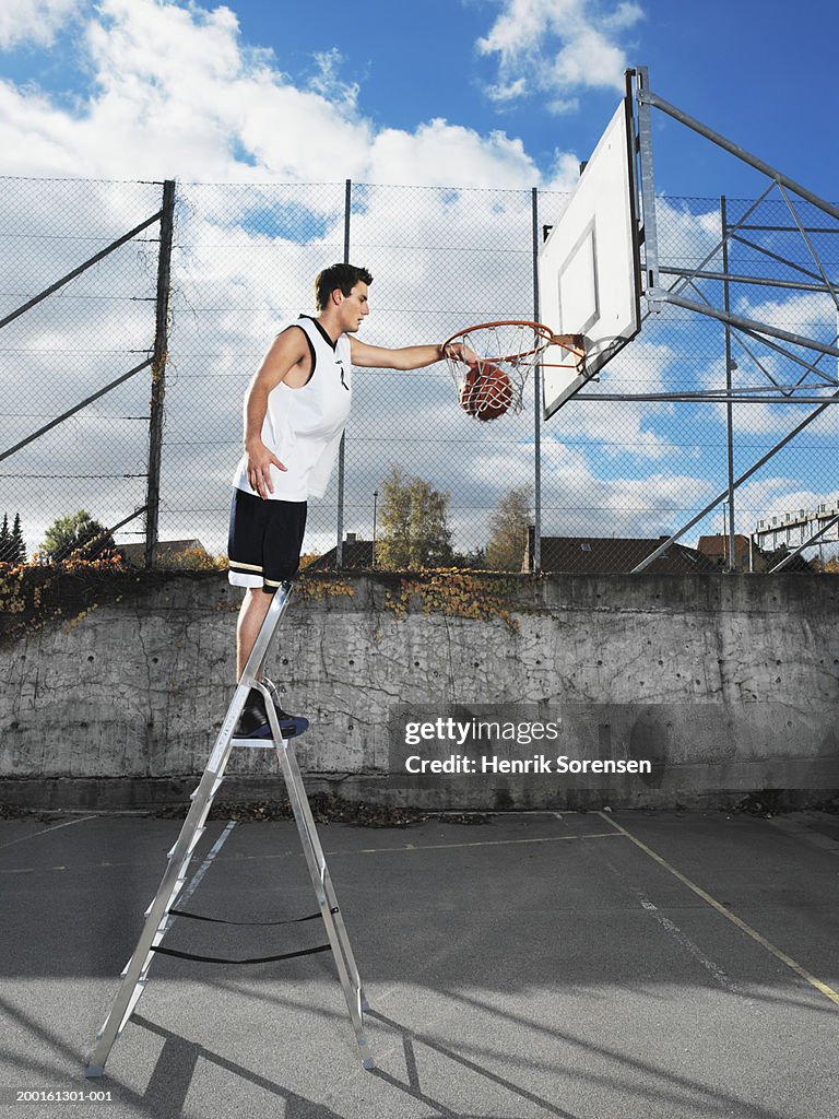 Young man on step ladder, dunking basketball in hoop, side view