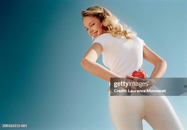 young woman with apple behind back, smiling, portrait, low angle view - white pants stock-fotos und bilder