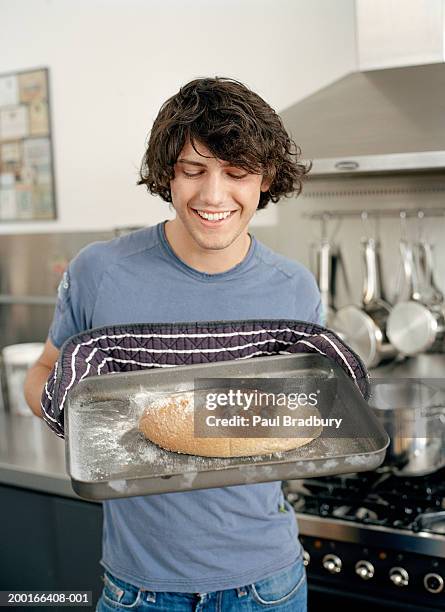 young man wearing oven gloves, holding tray with bread roll, smiling - baking tray stock pictures, royalty-free photos & images