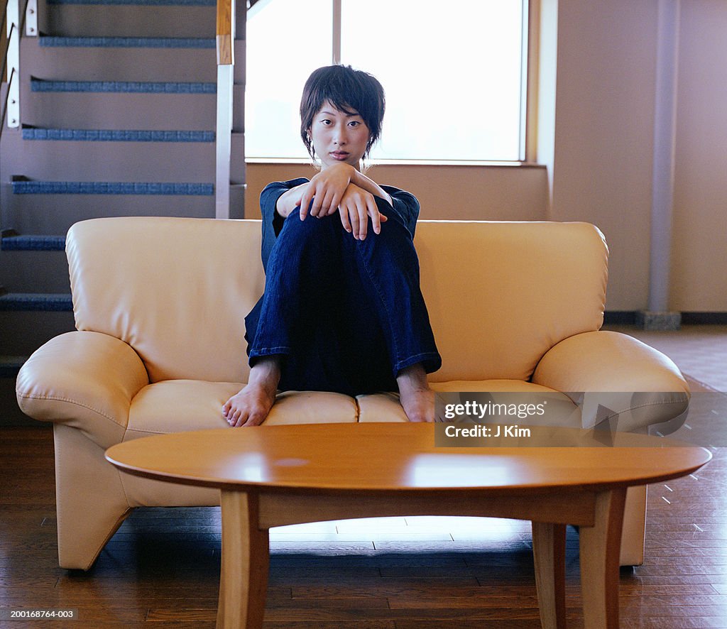 Young woman sitting on sofa, resting arms on knees, portrait