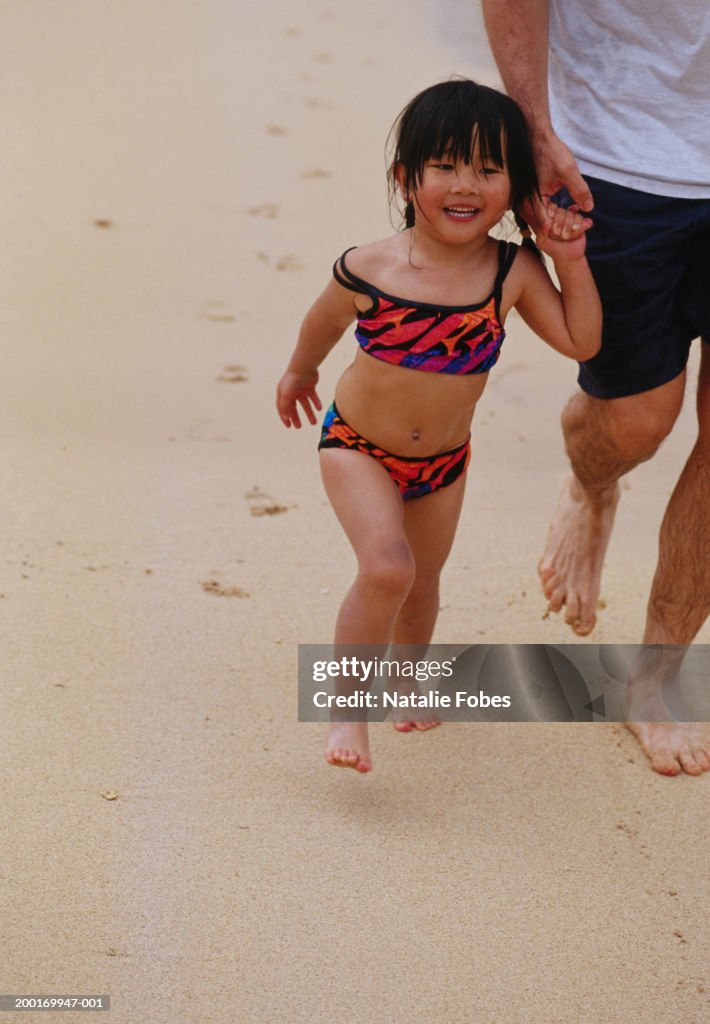 Girl (2-4) wearing swimsuit, holding father's hand on beach, smiling