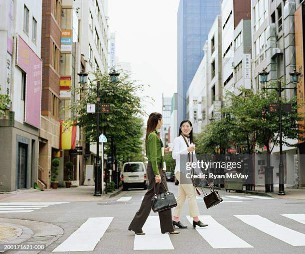 two young women crossing urban street, having conversation, side view - woman walking side view stock pictures, royalty-free photos & images