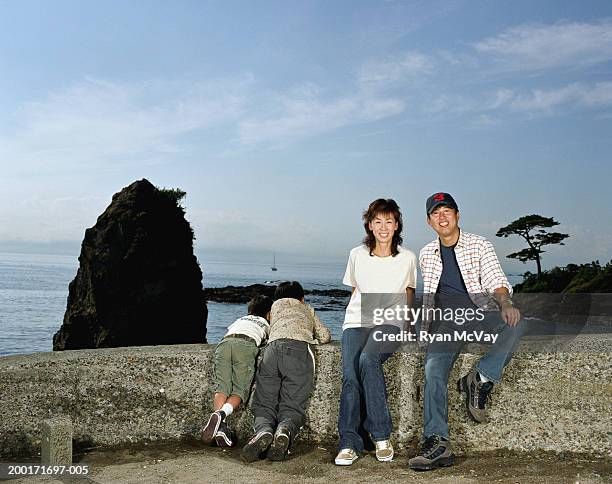 parents and two sons (5-10) on rock wall beside sea, parents smiling - front view portrait of four children sitting on rock stock-fotos und bilder