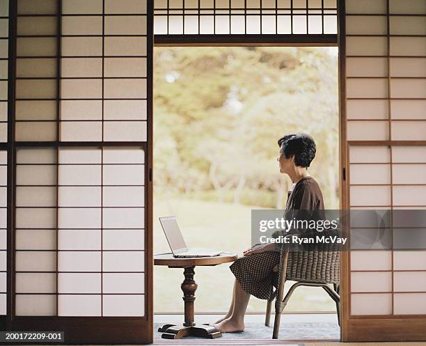 senior woman sitting beside open doorway, using laptop, side view - porta di carta giapponese foto e immagini stock