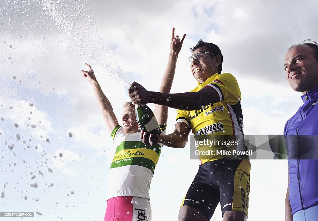 Three male cyclist standing on winner's podium, one opening champagne