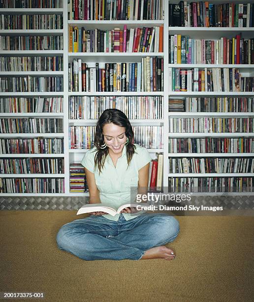 woman sitting cross legged, reading book, bookshelves in background - rd 1 stock-fotos und bilder